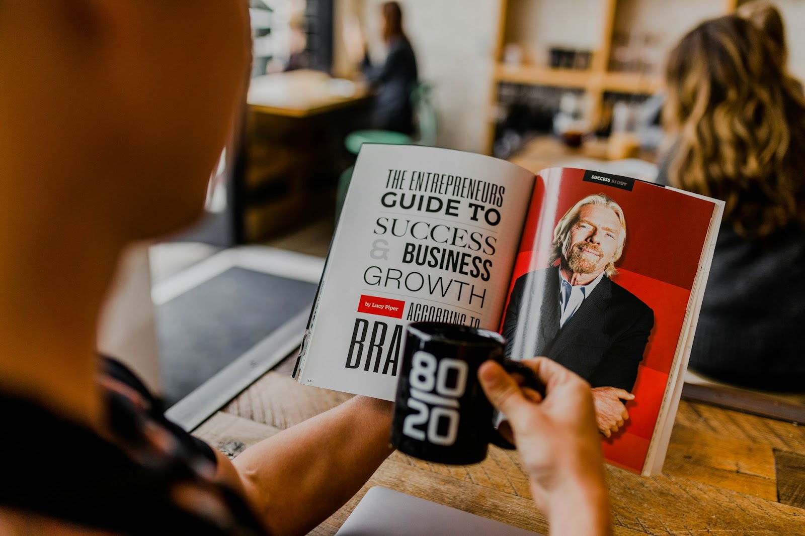 A person holds a black mug with "80 20" printed on it while reading a magazine featuring an article titled "The Entrepreneurs Guide to Success & Business Growth.