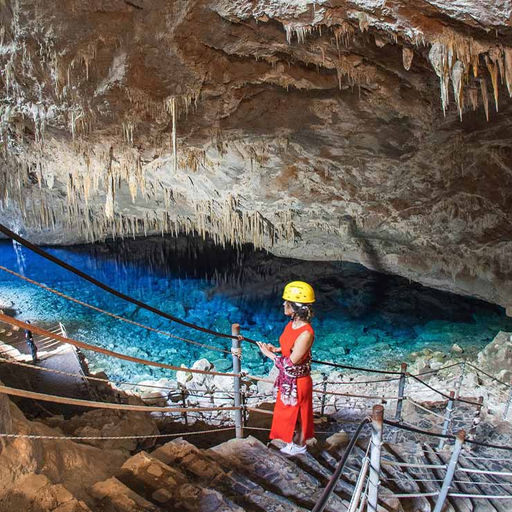 Pessoa usando capacete amarelo, observando uma lagoa azul dentro de uma caverna com estalactites penduradas no teto