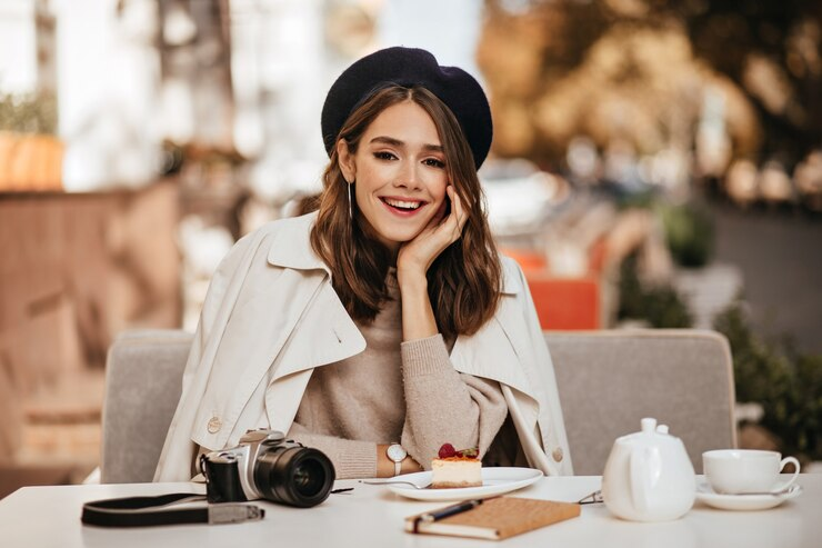 Smiling woman with brunette wavy hairstyle, beret, beige trench coat having lunch at a cafe