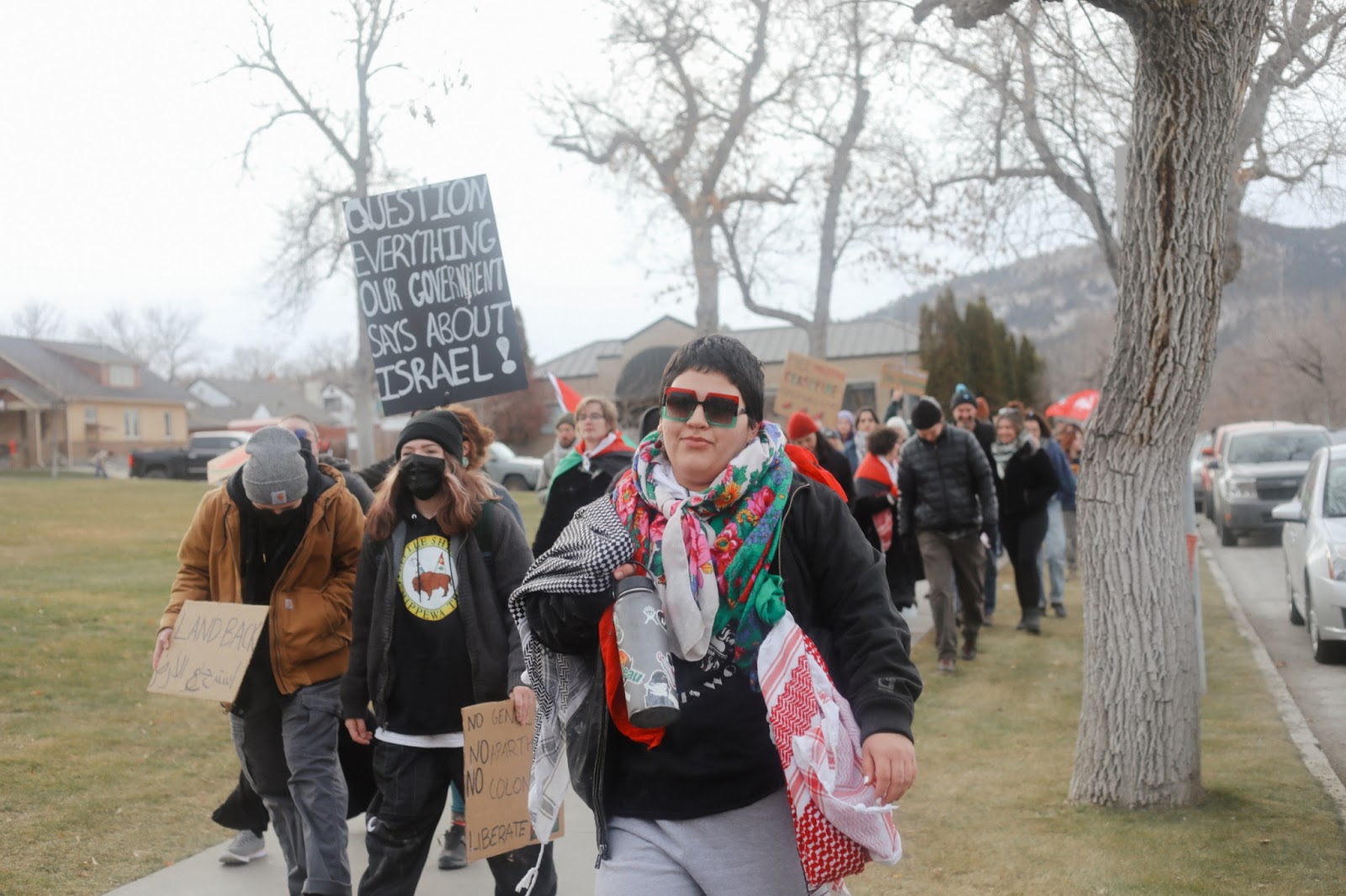 Color photograph of a person wearing green- and red-rimmed sunglasses and multiple keffiyehs leading a pro-Palestine protest down a suburban sidewalk