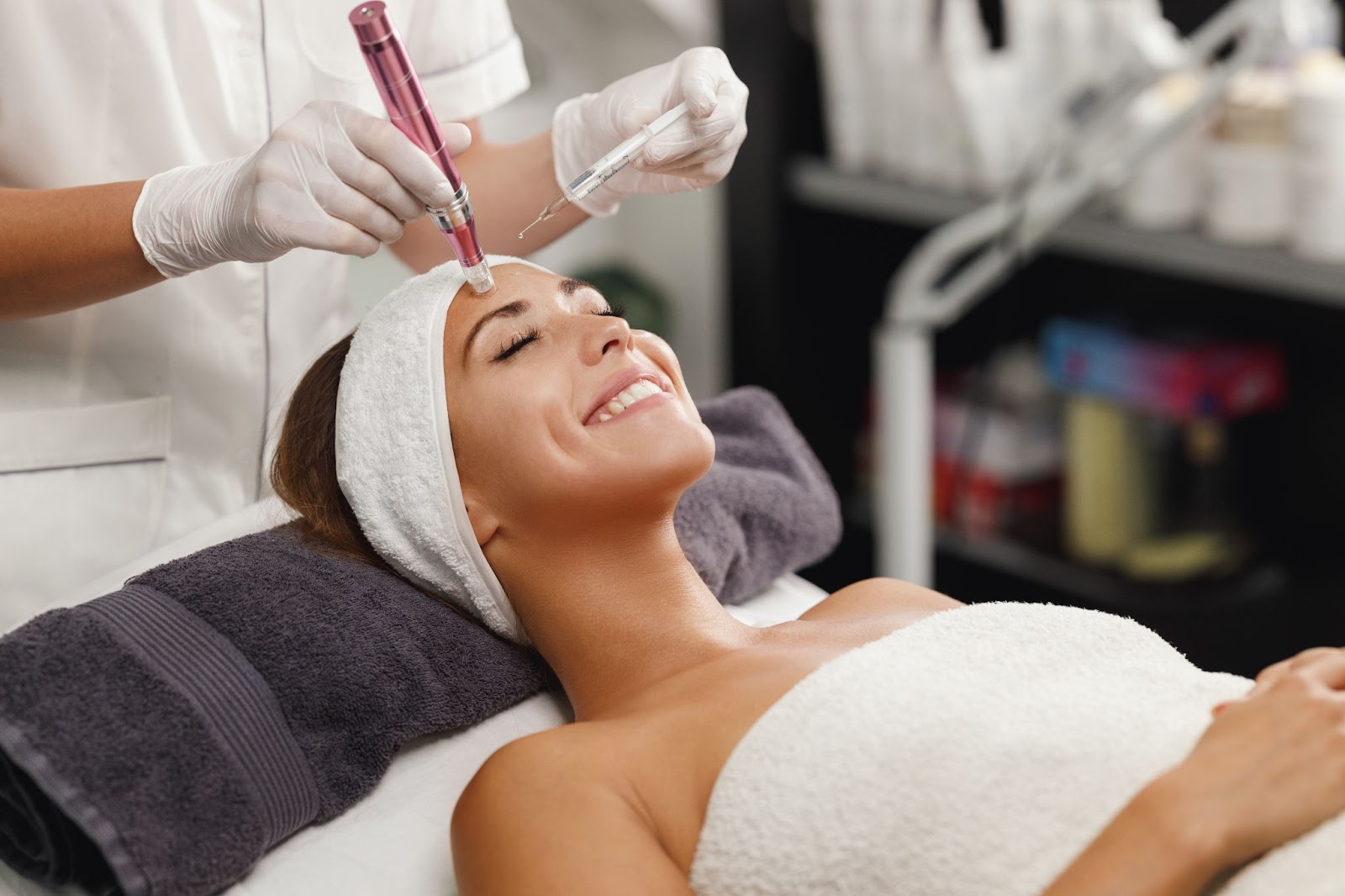 A smiling young woman relaxing on a table while an aesthetician applies microneedling treatment.