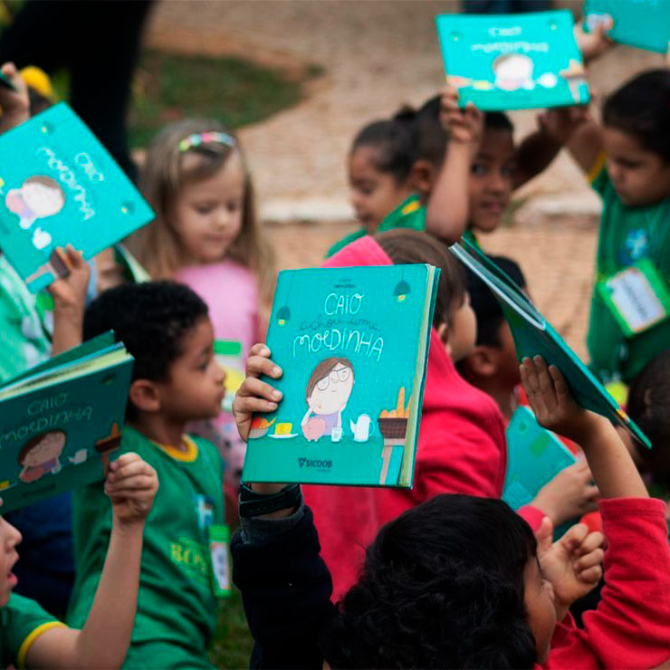 Crianças segurando e mostrando livros "Caio achou uma moedinha" durante um evento literário ao ar livre.