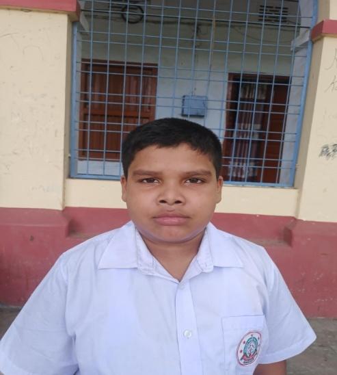 South Asian teen boy with short hair standing outside a school hallway in front of a window. He's in a white collared school uniform shirt. 