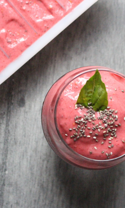 Smoothie rose aux fruits rouges et graines de chia, garni d'une feuille de basilic, servi dans un verre transparent sur un fond gris.