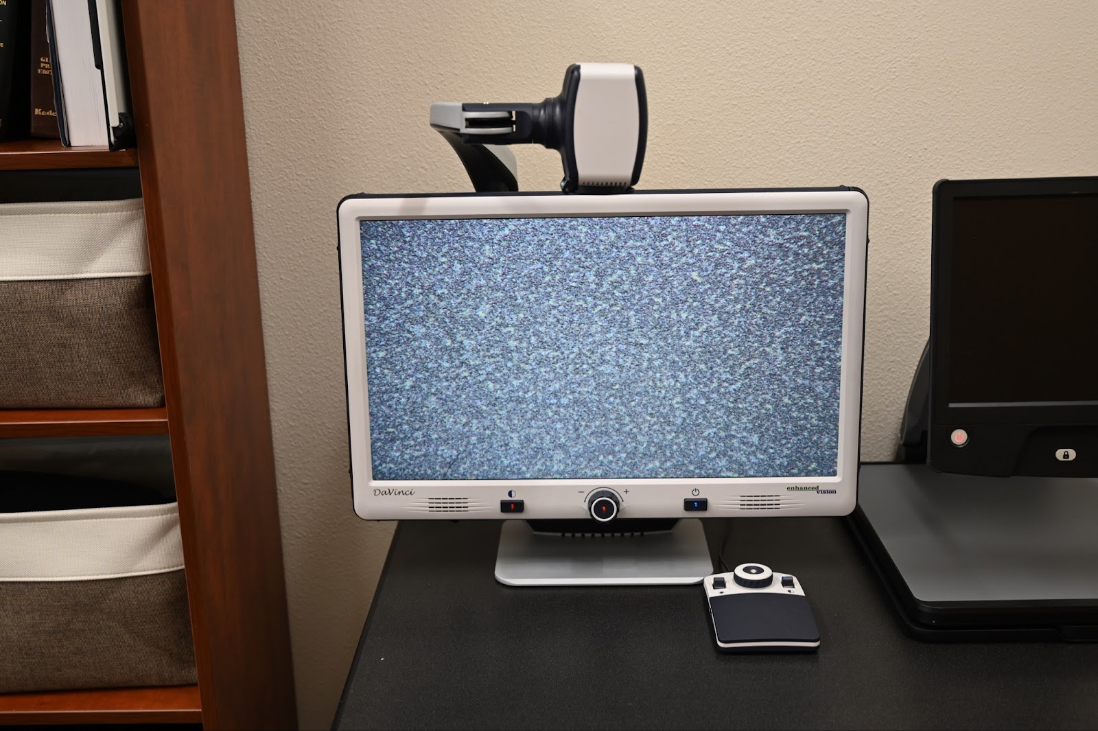 The image shows a desktop electronic magnifier sitting on a black desk. The magnifier features a large screen displaying static noise. Above the screen, there's a mounted camera. 