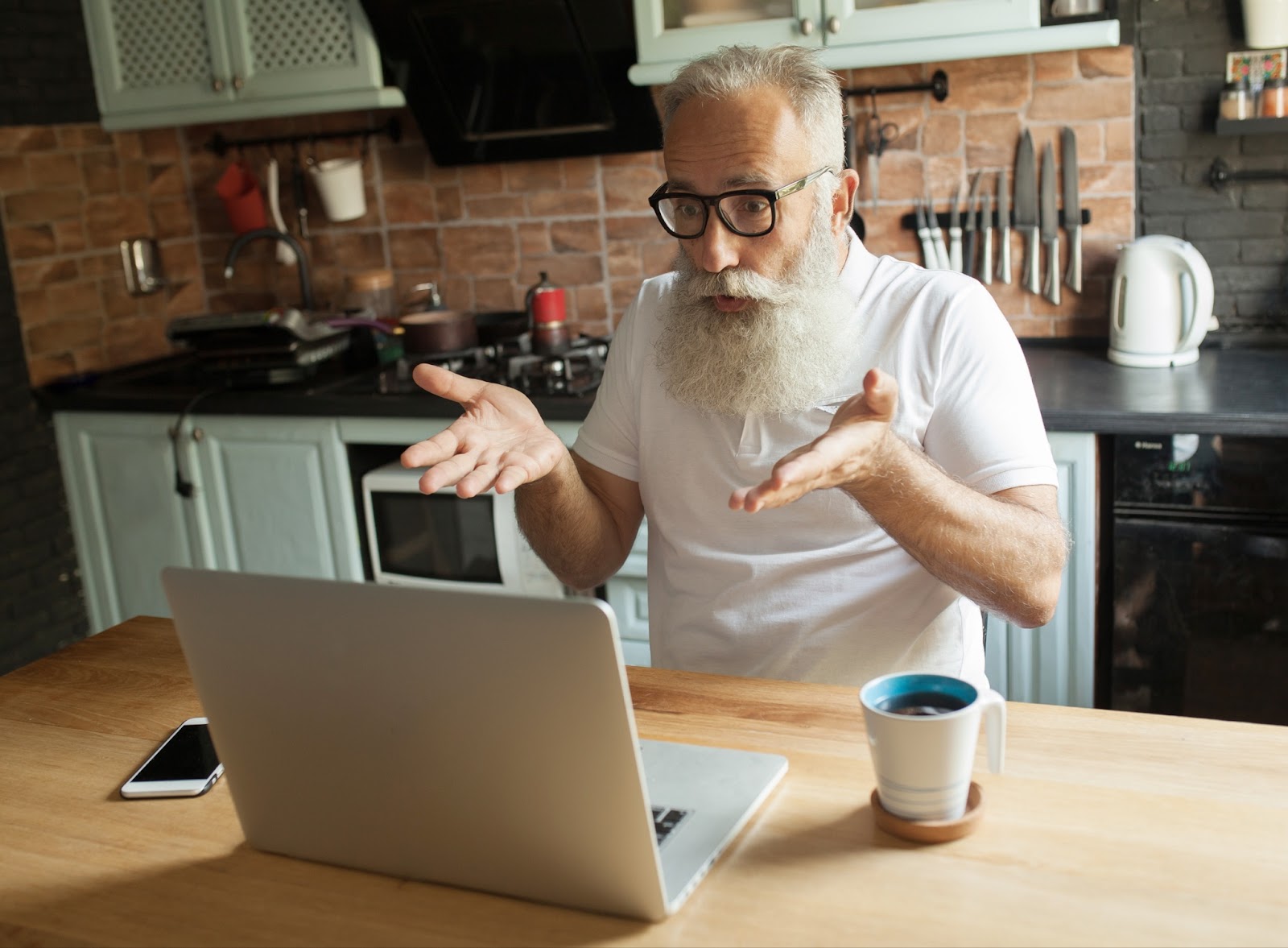 Caucasisan senior man gesturing frustratedly at his open laptop in the kitchen.
