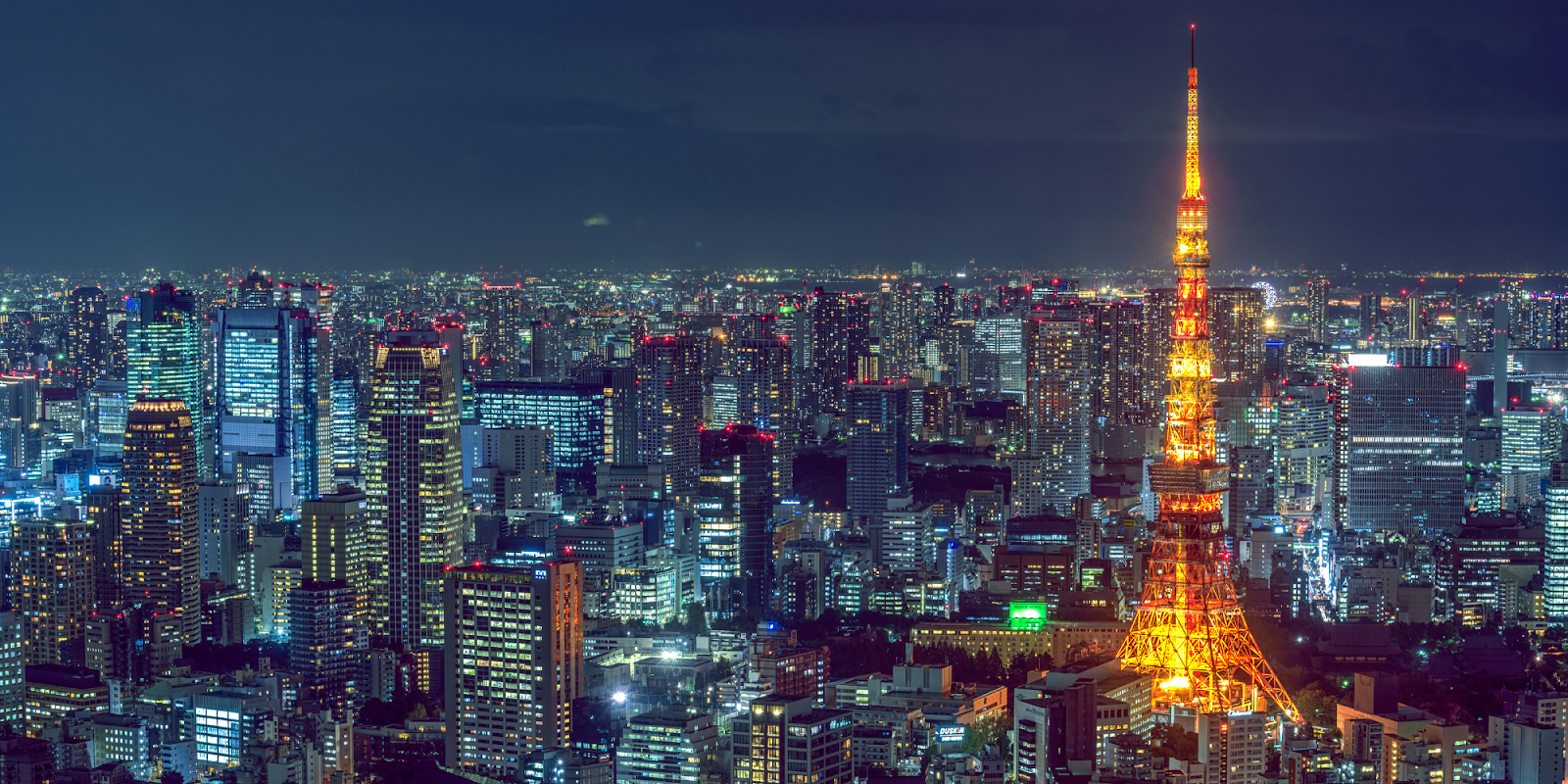 A nighttime panorama of Tokyo, Japan, with the Tokyo Tower brightly illuminated in the foreground. The cityscape is filled with countless skyscrapers and buildings, all aglow with lights, creating a mesmerizing and vibrant scene. The dense urban environment stretches into the horizon, showcasing Tokyo's bustling and dynamic nature.