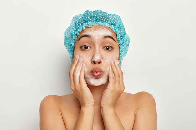 Close up portrait of lovely female with folded lips, applies washing soap on face, washes skin for looking fresh and clean, stands with naked body, pampers face