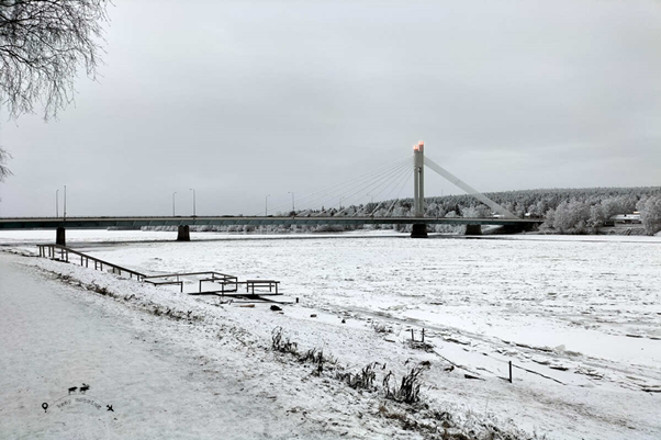 The photograph of the most famous bridge in Rovaniemi. You can see the frozen river covered in snow and the arctic forest beyond the river.