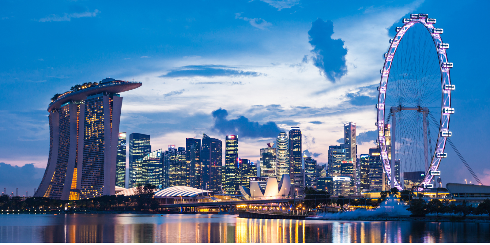 A breathtaking view of Singapore's Marina Bay at dusk, featuring the iconic Marina Bay Sands hotel with its unique architecture on the left and the Singapore Flyer Ferris wheel on the right. The skyline is adorned with modern skyscrapers, all brightly lit, reflecting on the tranquil waters of the bay. The scene captures the elegance and futuristic charm of Singapore as the evening sets in.