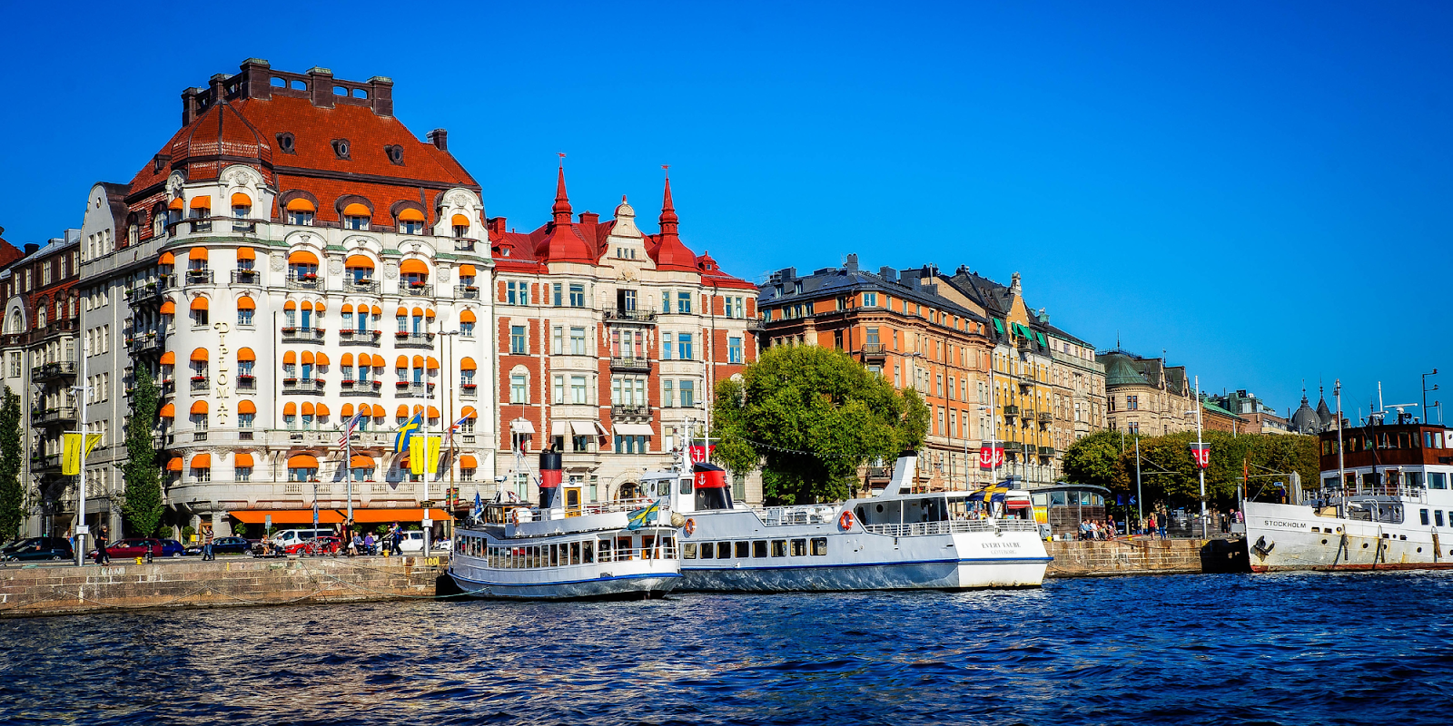 A vibrant waterfront scene in Stockholm, showcasing beautiful historic buildings with colorful facades and red roofs. Boats, including accessible travel options, are docked along the quay, and Swedish flags wave in the breeze. The clear blue sky adds to the picturesque atmosphere of the city.