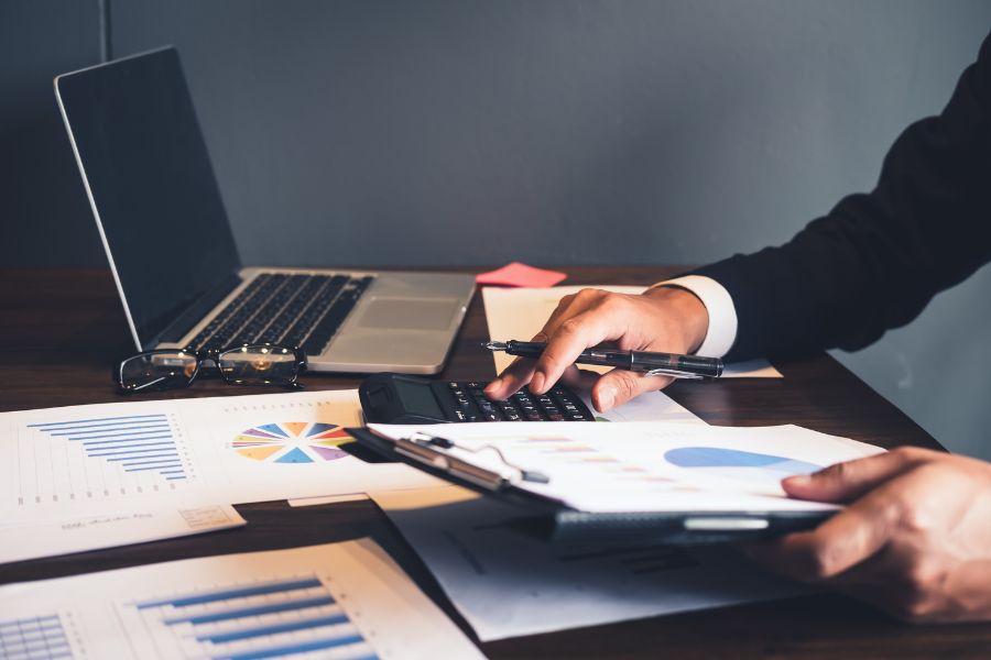 Person using calculator with financial reports and laptop on desk.