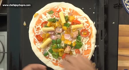 A rolled-out pizza dough on a baking sheet, spread with tomato sauce and topped with cheese and various vegetables.