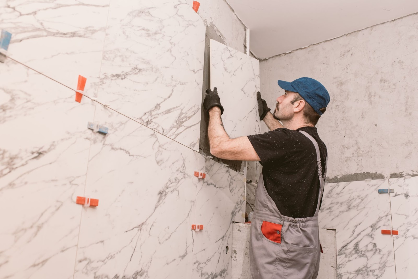  A remodeling contractor in a dark brown shirt, khaki overalls, blue cap, and black gloves installs ceramic tiles, marking them with orange and blue tapes inside a bathroom.