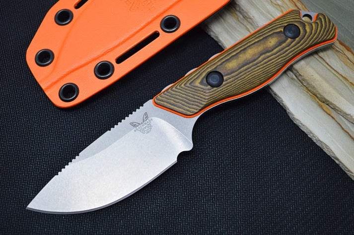 An image featuring the Benchmade 15017-1 Hidden Canyon Hunter on a black and wooden background. The Benchmade logo can be prominently seen on the knife's blade. Next to the knife is a bright orange sheath.