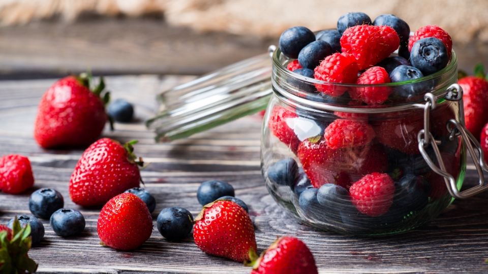 A glass jar filled with fresh berries, including strawberries, raspberries, and blueberries, sits on a rustic wooden table.