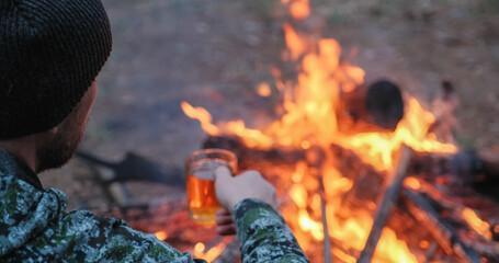 Man with cup of tea the fire in the forest. Human looks at the bonfire and drinks tea on the nature in the summer. Alone traveler sits and rests by the fire with a cup of coffee at dusk.