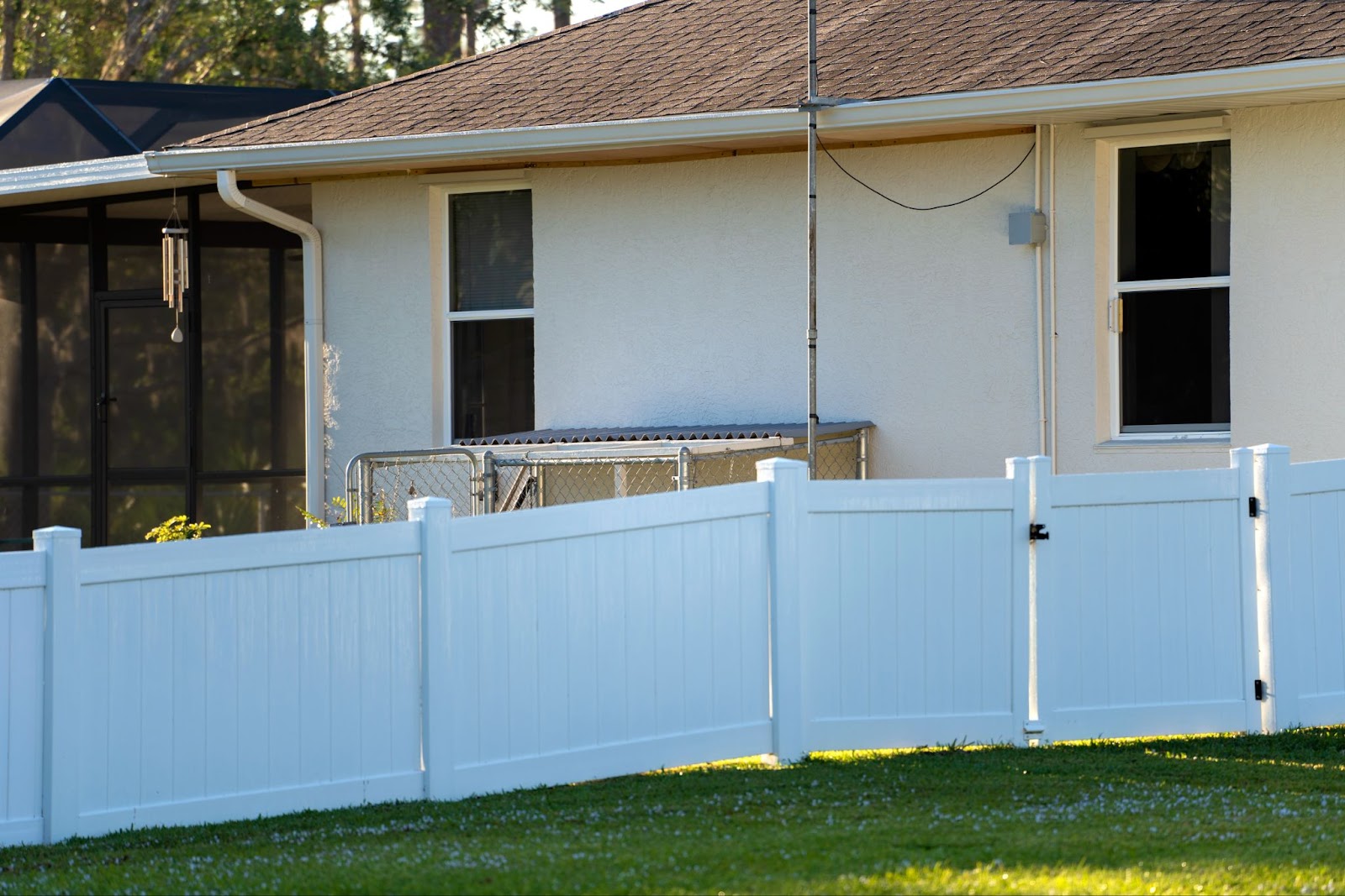 A white vinyl picket fence encloses a house from the backyard's lush green lawn, defining the property's boundaries.