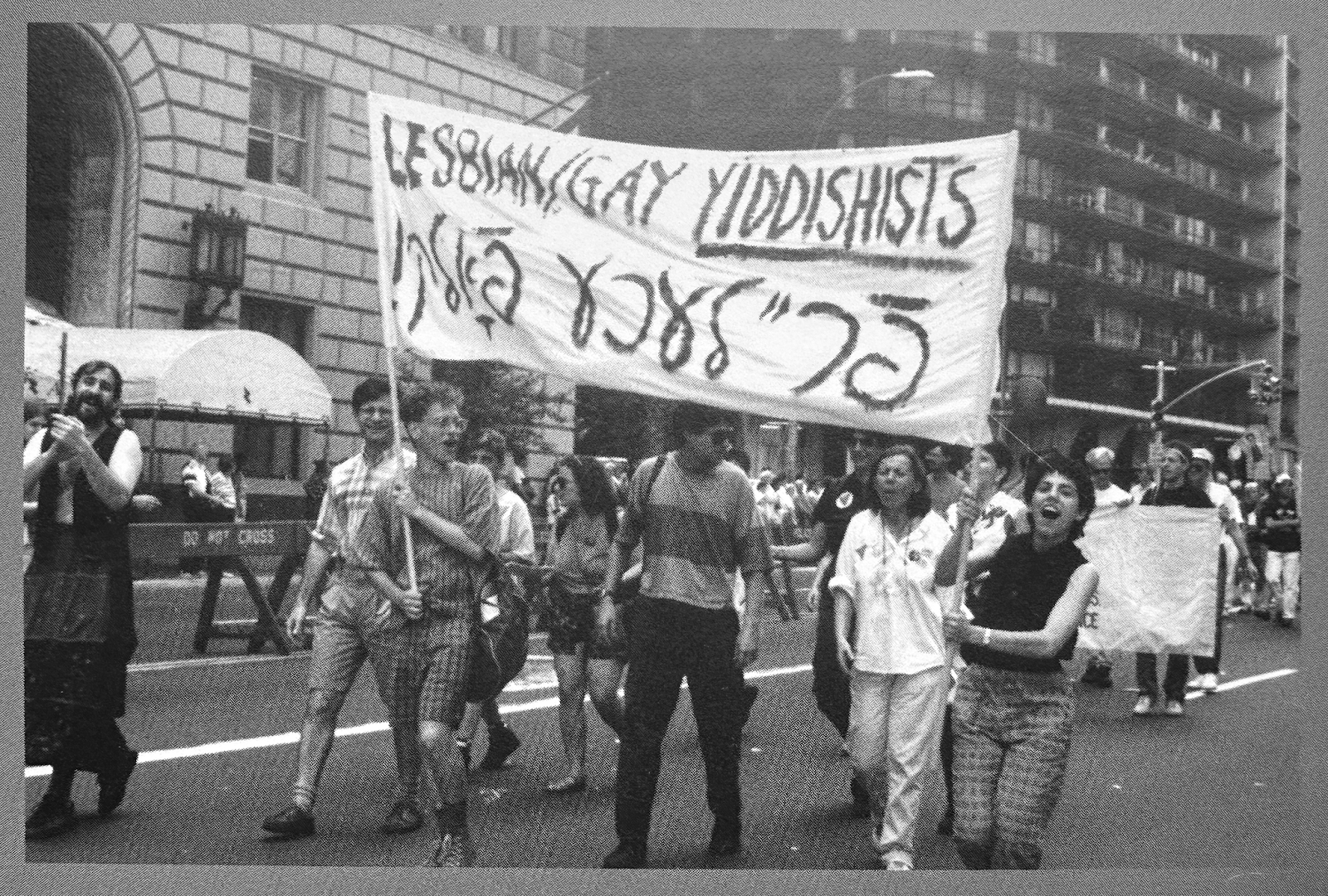 Black and white image of 1989 Gay Pride March in NYC. Yiddish banner reads: “Gay people!” Irena Klepfisz is 2nd from right, in white.