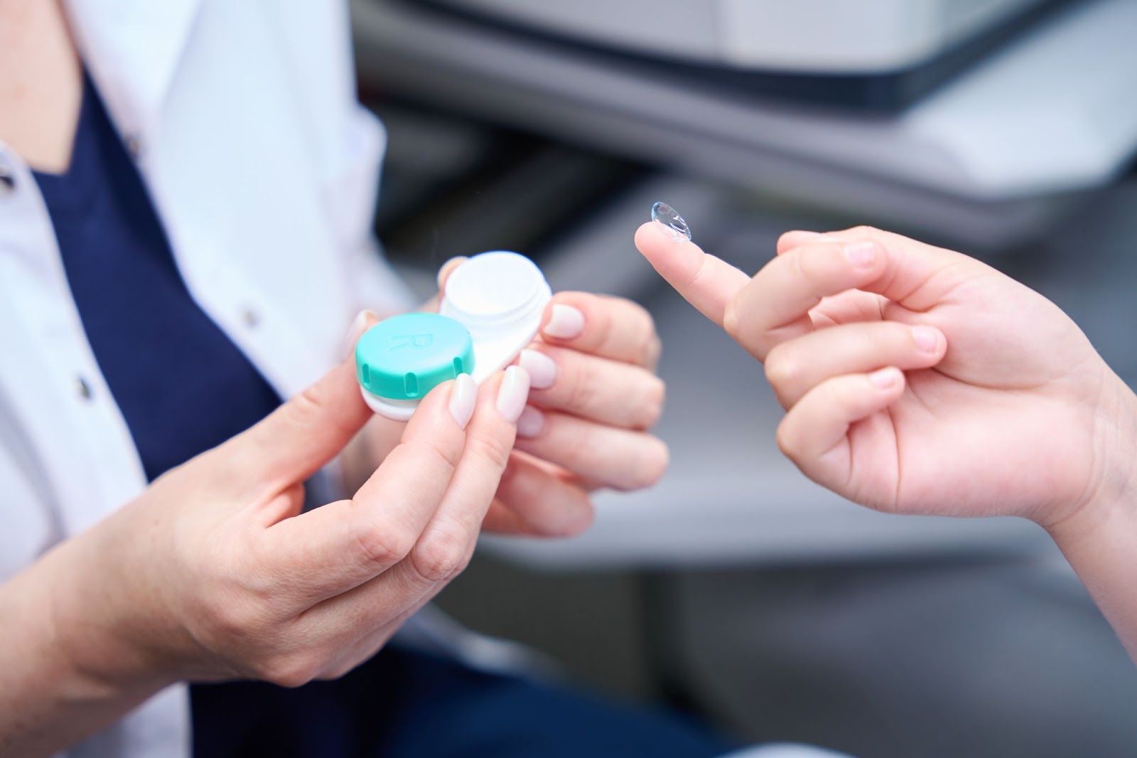 An optometrist holds a contact lens case while her patient practices inserting a contact lens with her right index finger.