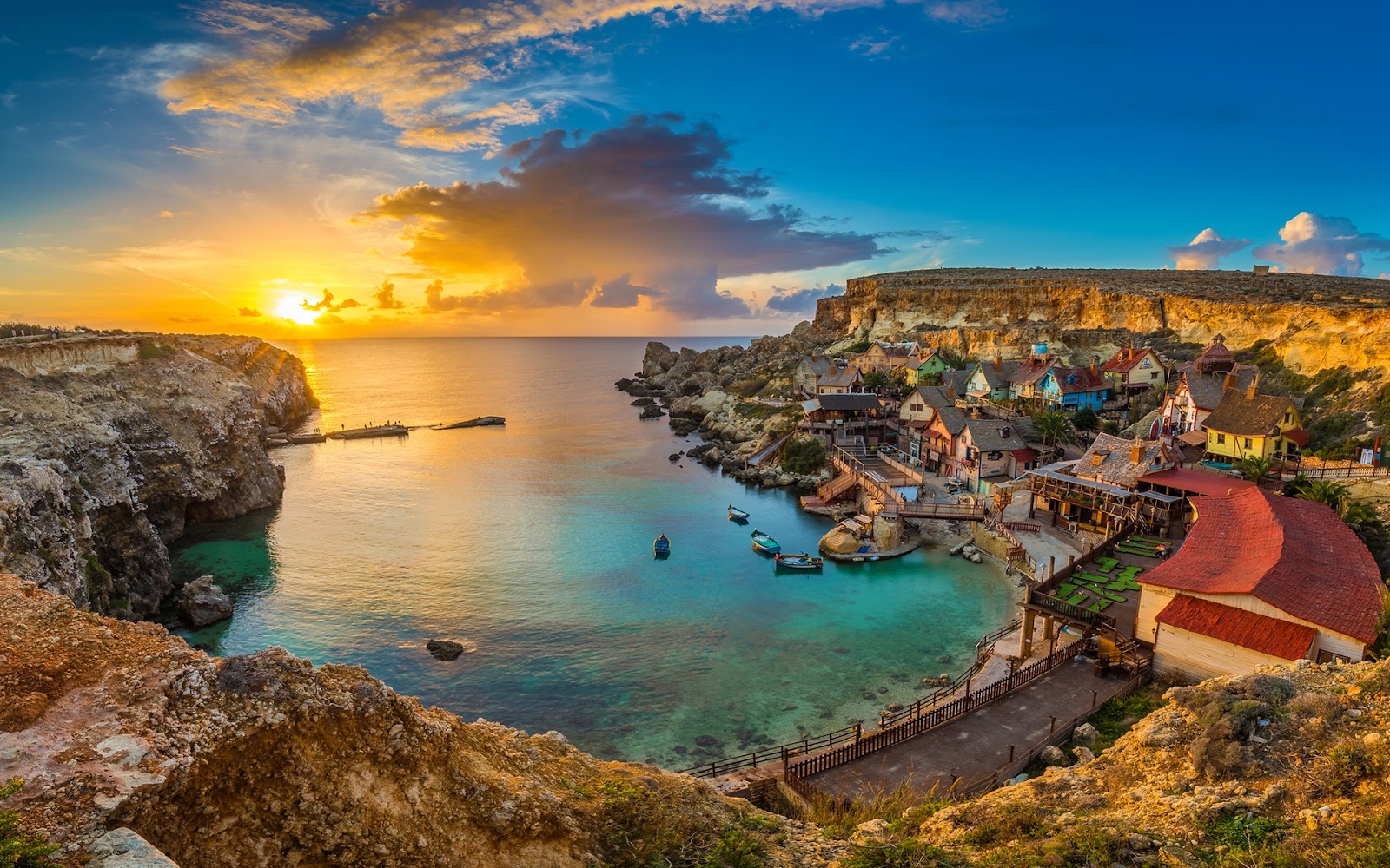 Sunset over water with silhouettes of rocks and houses against the colorful sky.