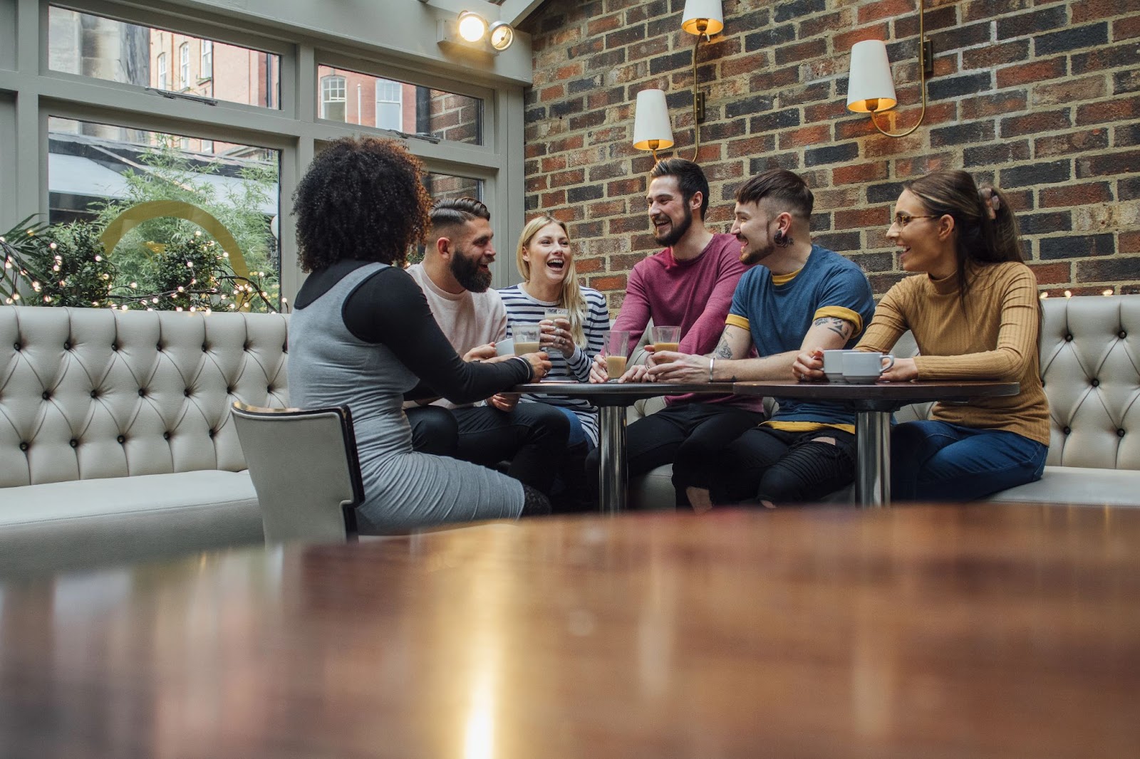 A group of six people sit around a table in a cozy, brick-walled café, engaged in conversation and laughing together, enjoying the warmth of in-person meetings.