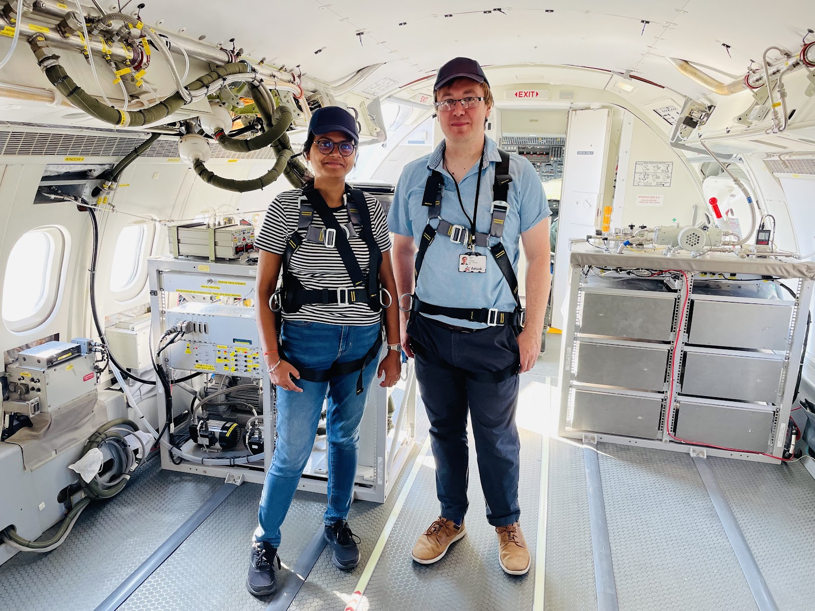 Two people in safety harnesses, standing in front of a rack of scientific equipment in the cabin of a research aircraft.