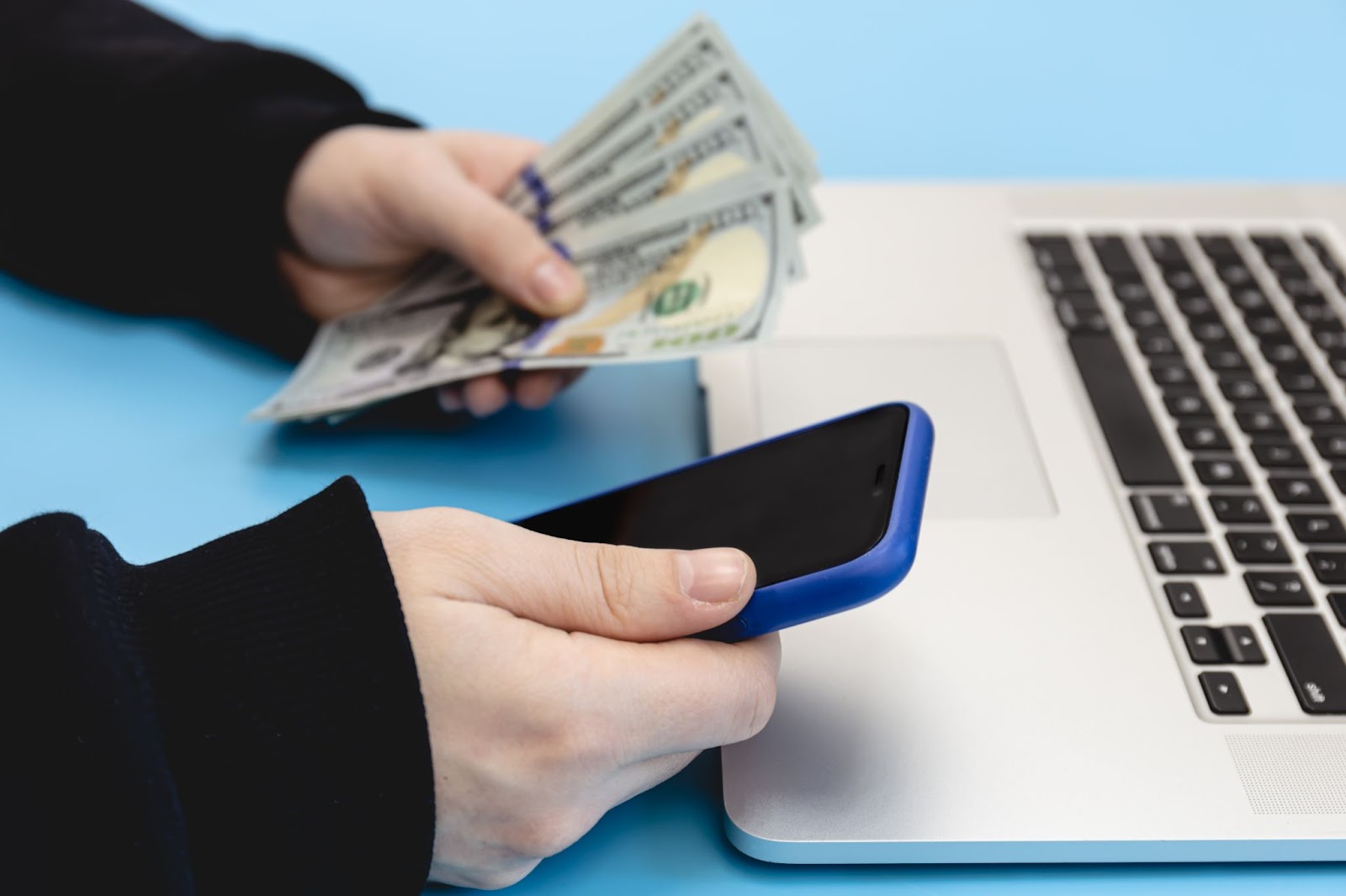 Hands holding cash and a smartphone over a laptop, suggesting online financial transaction or digital payment