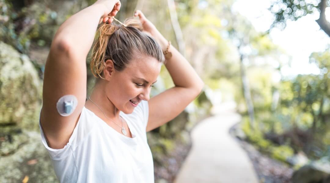 A young woman outdoors tying her hair back, wearing a continuous glucose monitor on her upper arm.