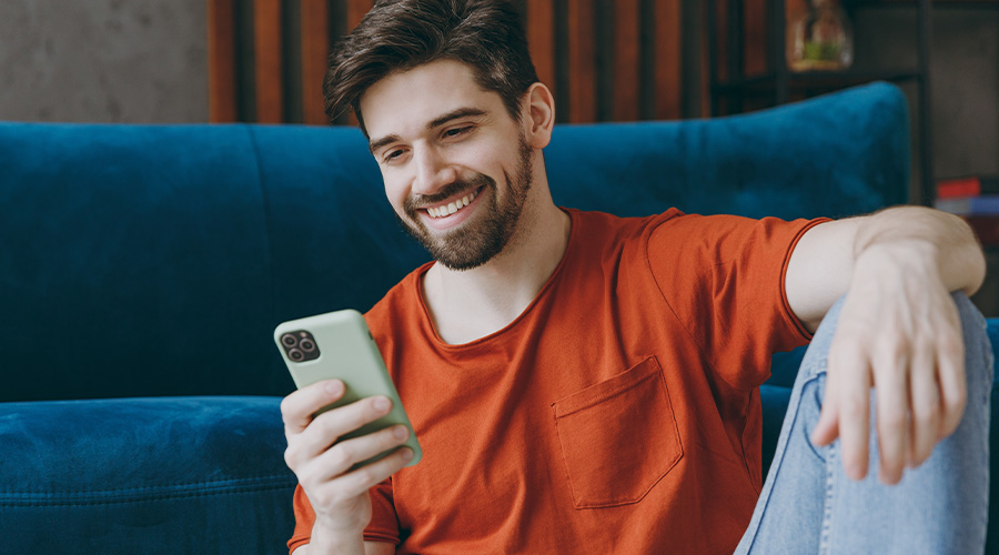 Brunette man sitting on the floor and leaning against a blue couch while smiling and looking at his phone.