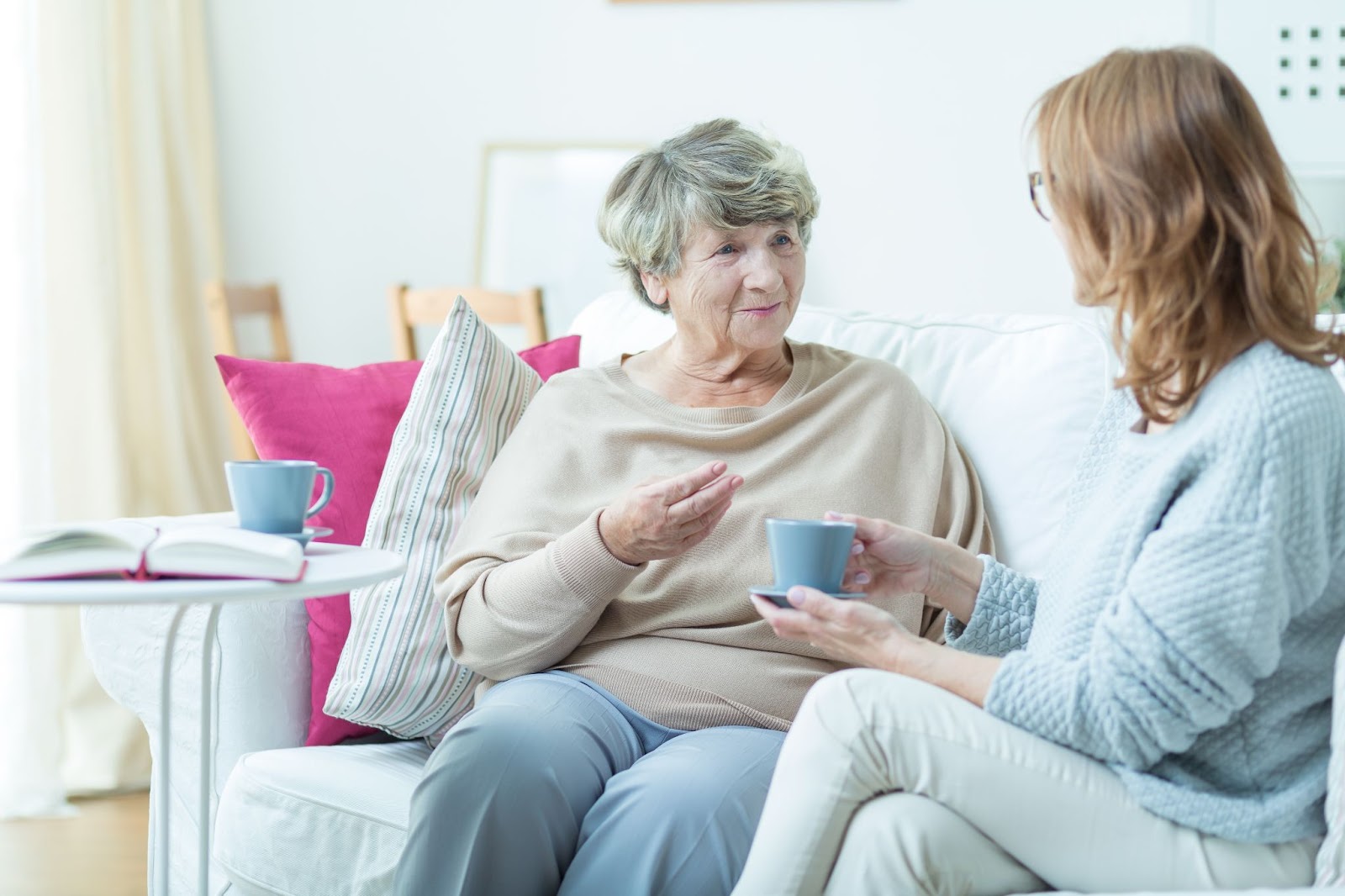 A senior woman and her daughter sitting on a couch drinking tea and talking about memory care.