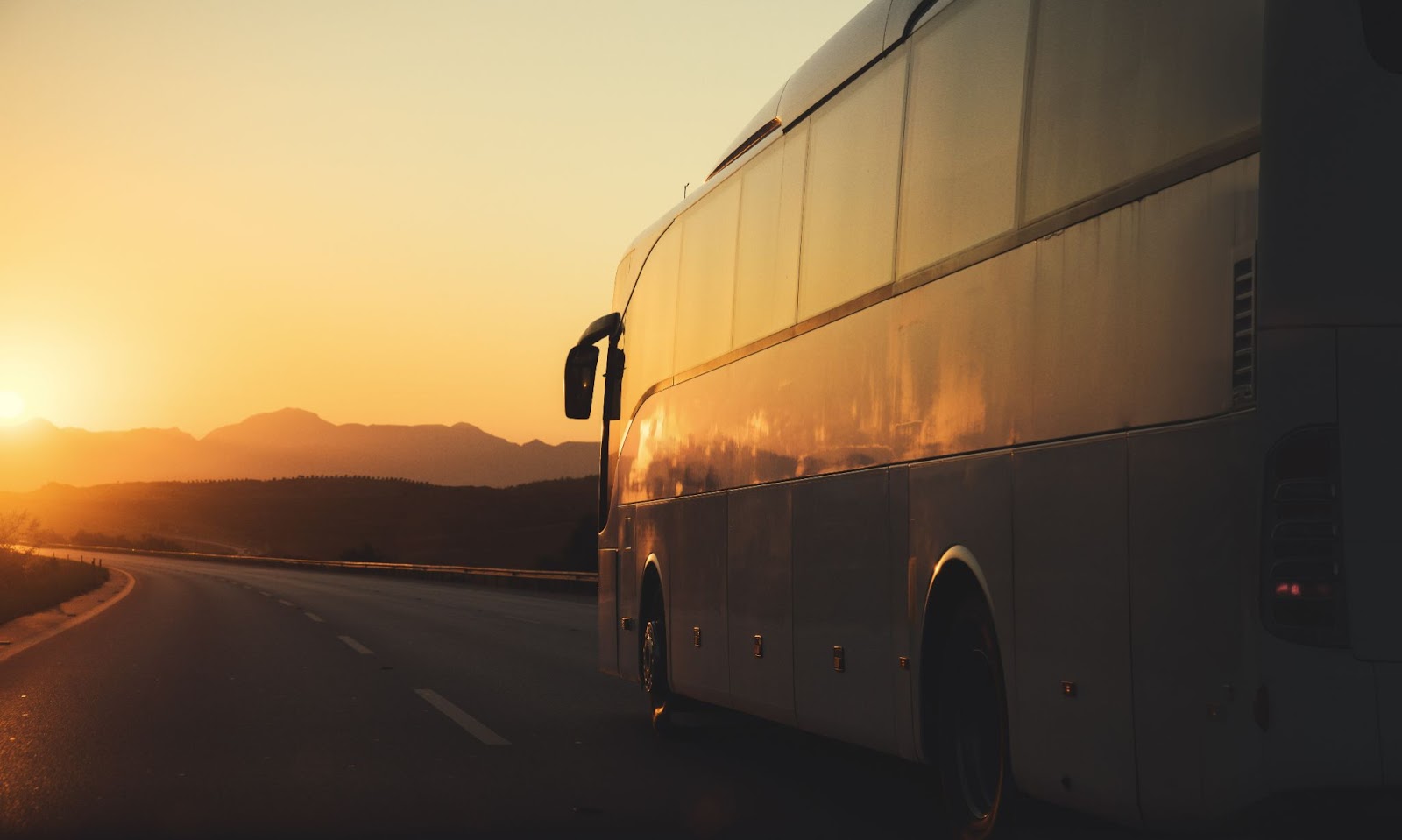Ônibus de viagem branco na estrada vazia, refletindo os tons alaranjados do pôr do sol ao horizonte.