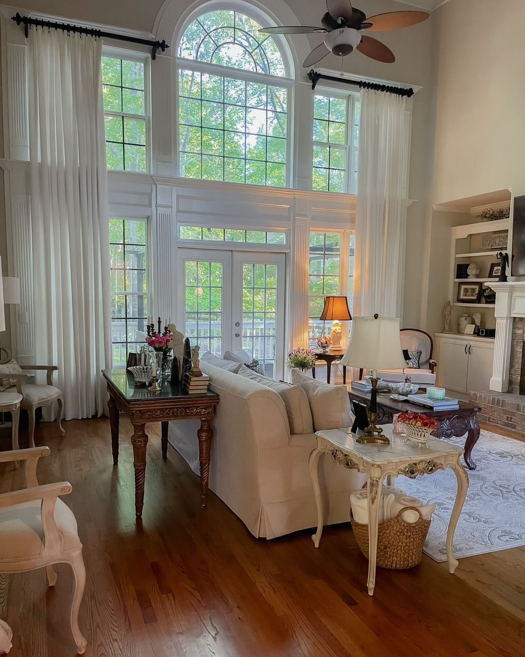 Spacious living room with tall arched windows & white drapes. Beige sofa with side table & lamp. Wooden table with decor. Cozy armchair by window. Fireplace with shelves. Mix of vintage & classical furniture.