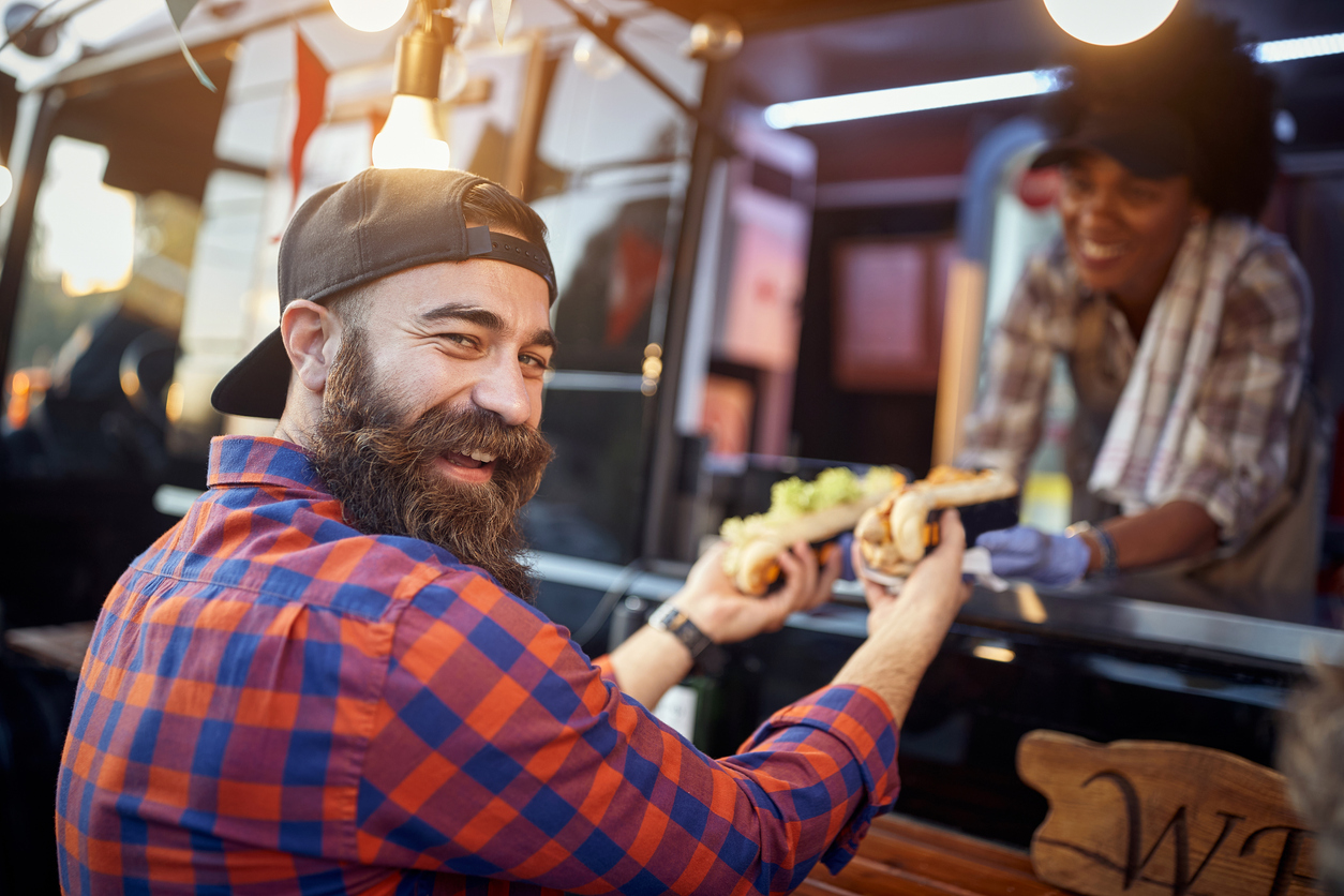 A man with a beard receiving hot dogs from a friendly employee at a food truck.