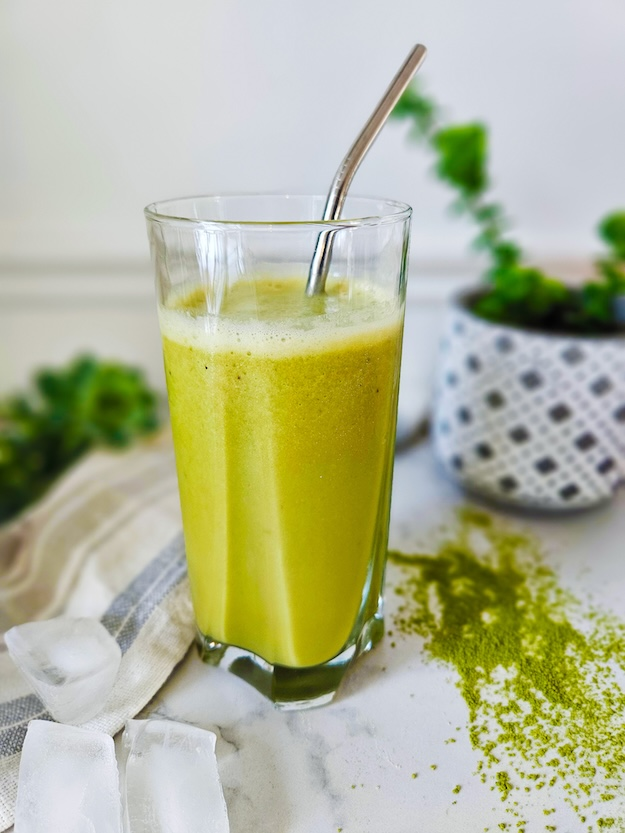 Energizing green smoothie served in a clear glass on a marble table, next to ice cubes and matcha powder.