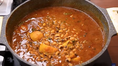 Boiled lobia and fried potatoes being mixed into the masala in a pan, creating a hearty and flavorful Lobia Aloo Masala ready to simmer.