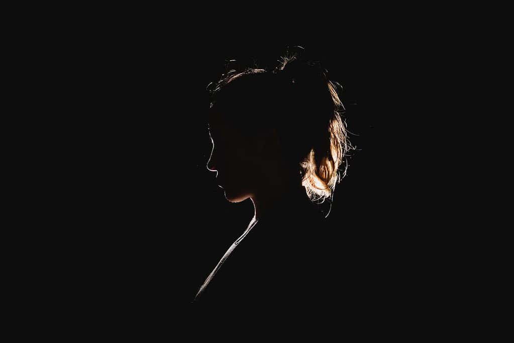 Silhouette of a person's profile against a dark background, with light outlining the shape of the hair, ear, and nose. The image captures a sense of mystery and minimalism due to the contrast between light and shadow.