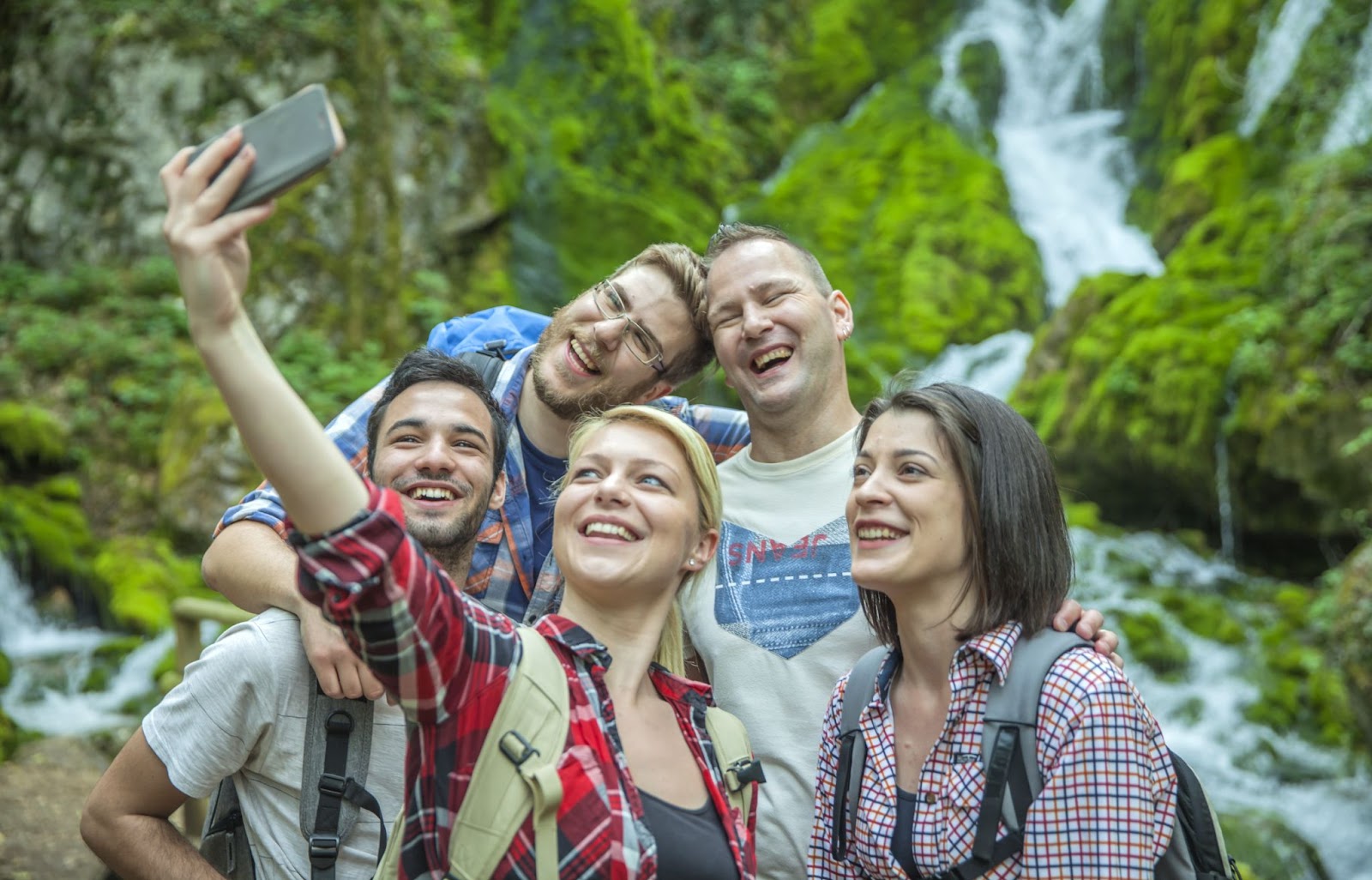 Imagem de cinco amigos fazendo uma selfie em meio a uma paisagem verde. Há duas mulheres e três homens na foto e todos sorriem para a tela do celular.