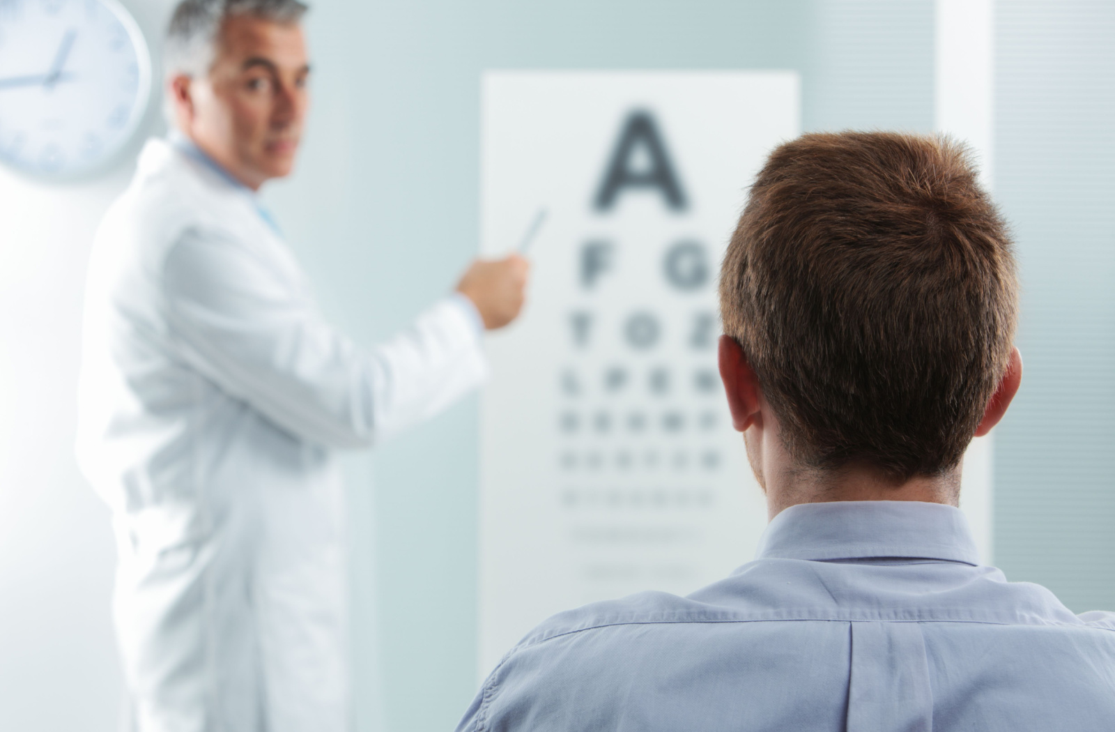 A man sits in front of a Snellen eye chart, and an optometrist is asking him to read the letters on the chart