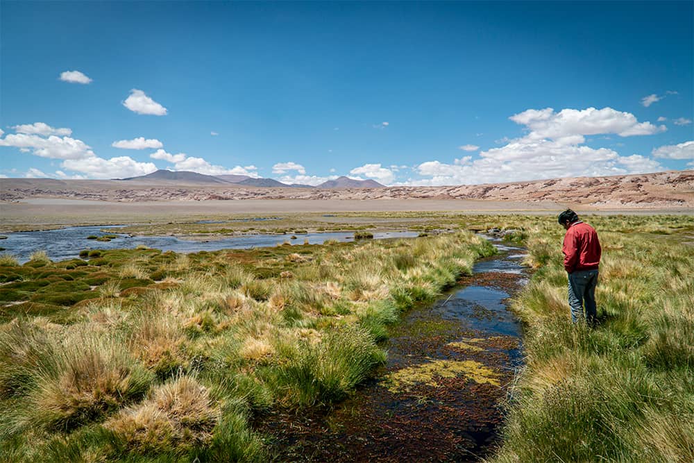 A man looks folorn at a river running through a mountainous wetland landscape