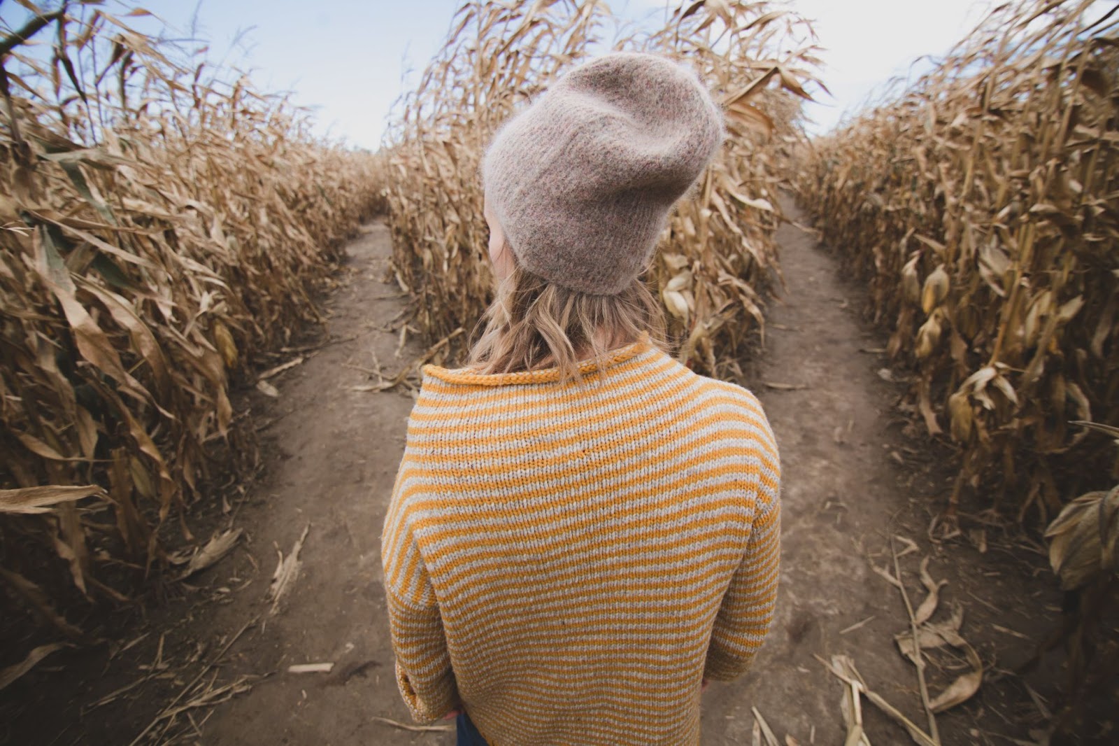 Woman in a sweater and a beanie staring at two paths in a corn maze.