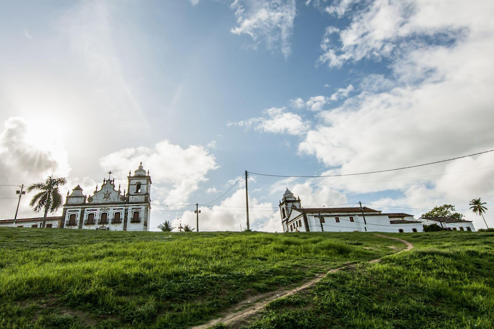 Morro de gramado com a Igreja de São Cosme e Damião no topo. Céu azul com grandes nuvens acinzentadas