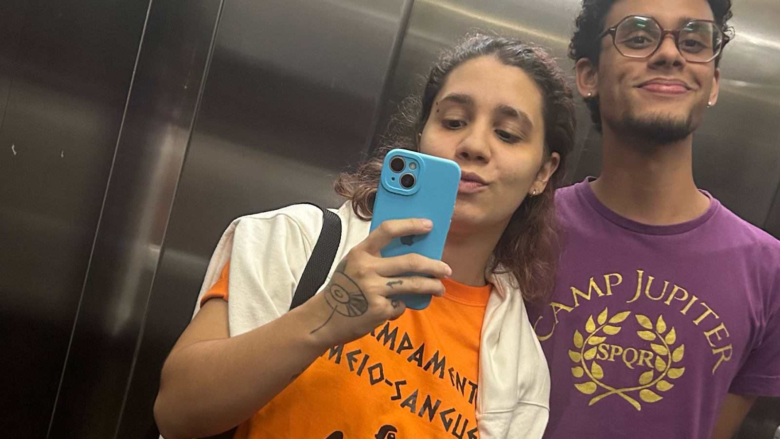 Iana Maciel, quem vos escreve, com a camiseta laranja do Acampamento Meio-Sangue, ao lado de seu amigo Victor Roldão, utilizando o uniforme roxo do Acampamento Júpiter. Fotografia retirada em um espelho de elevador.