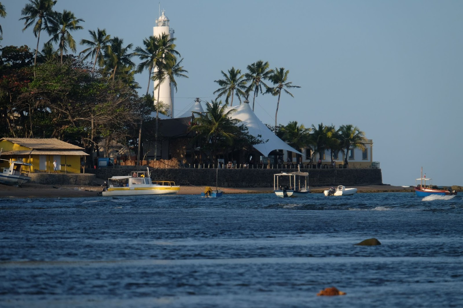 Mar sem ondas com barcos e lanchas próximos à areia em primeiro plano. Ao fundo da imagem, um estabelecimento com uma grande tenda branca e coqueiros cobrem parcialmente a vista do Forte.