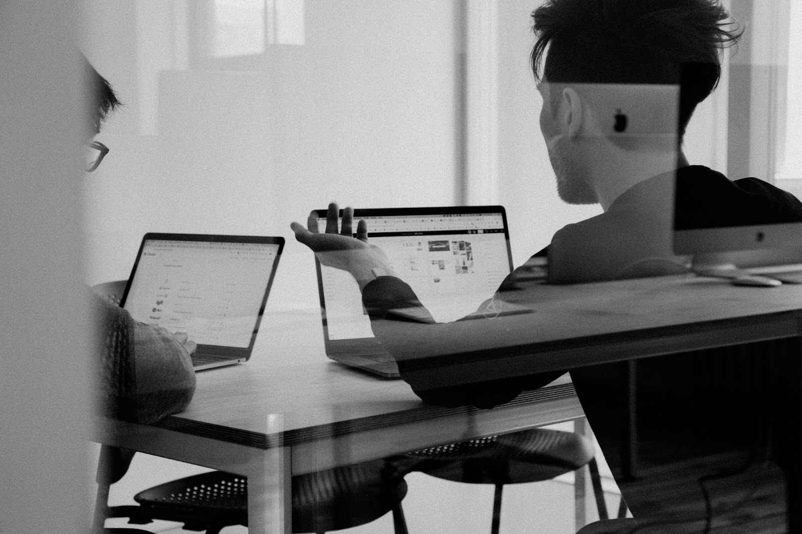 Pessoa sentada em uma mesa, trabalhando com dois laptops. O ambiente parece ser interno, iluminado pela luz natural que entra por uma janela. A pessoa está concentrada nas telas, envolvida em seu trabalho
