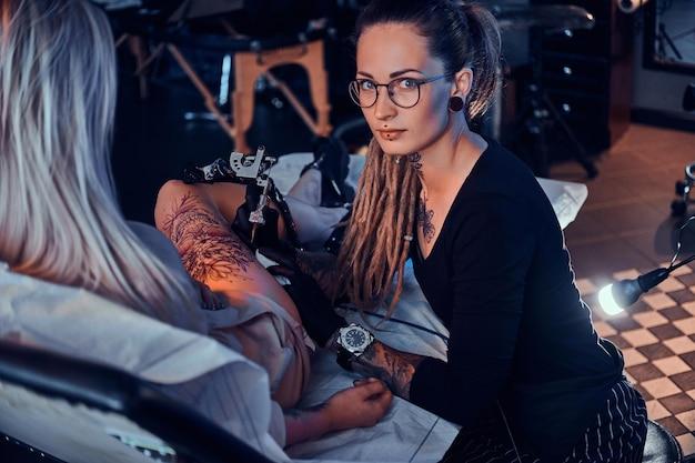 Beautiful creative master with dreadlocks is working on new peace of big leg tattoo for customer.