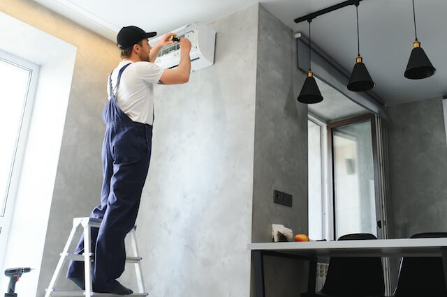An HVAC technician on a stepladder services an air conditioner in a modern kitchen, performing adjustments that help address and prevent issues like noise commonly caused by air ducts.