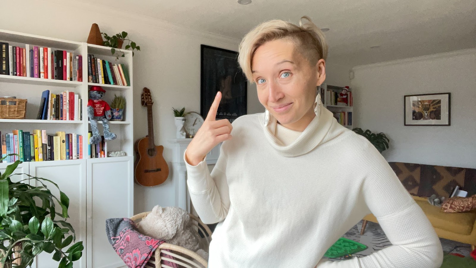 Email marketing expert Tarzan Kay stands in her living room with an amused expression, pointing to her cheek with one finger. She has short, partially shaved, white-blonde hair and wears a white turtleneck sweater. Behind her is a white bookshelf filled with books of various colors, a teddy bear in red attire, and small plants. To the left, a classical guitar hangs on the wall. There is also a dark framed picture on the wall, a green plant to the right, and in the foreground, a cushioned chair with a pink patterned blanket and a plush toy on it. The room is well-lit with natural light.