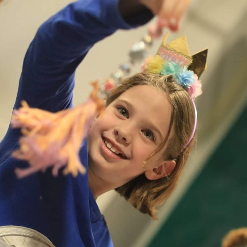 A young girl wearing a festive, party headband holds up a shiny object