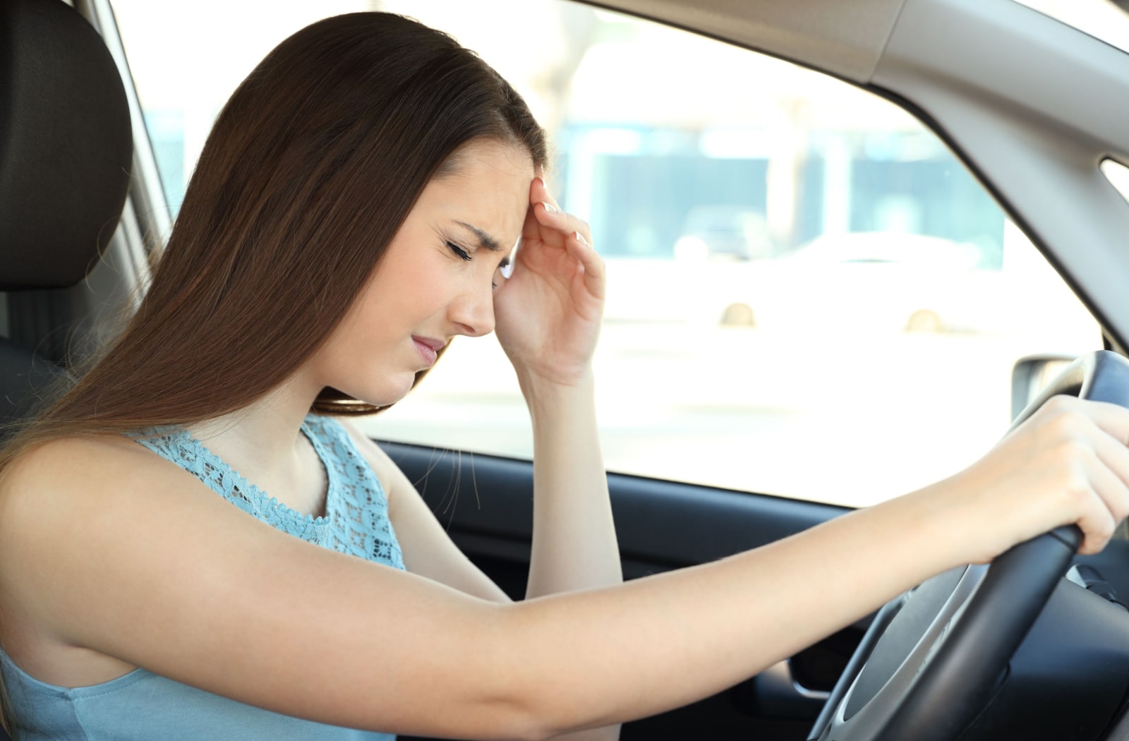 A woman driving and placing her left hand on her forehead, as she's feeling dizzy while driving
